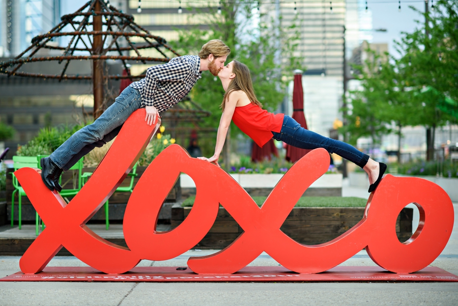 An engaged couple reach for a kiss on top of the xoxo statue outside of 30th street station in philadelphia.